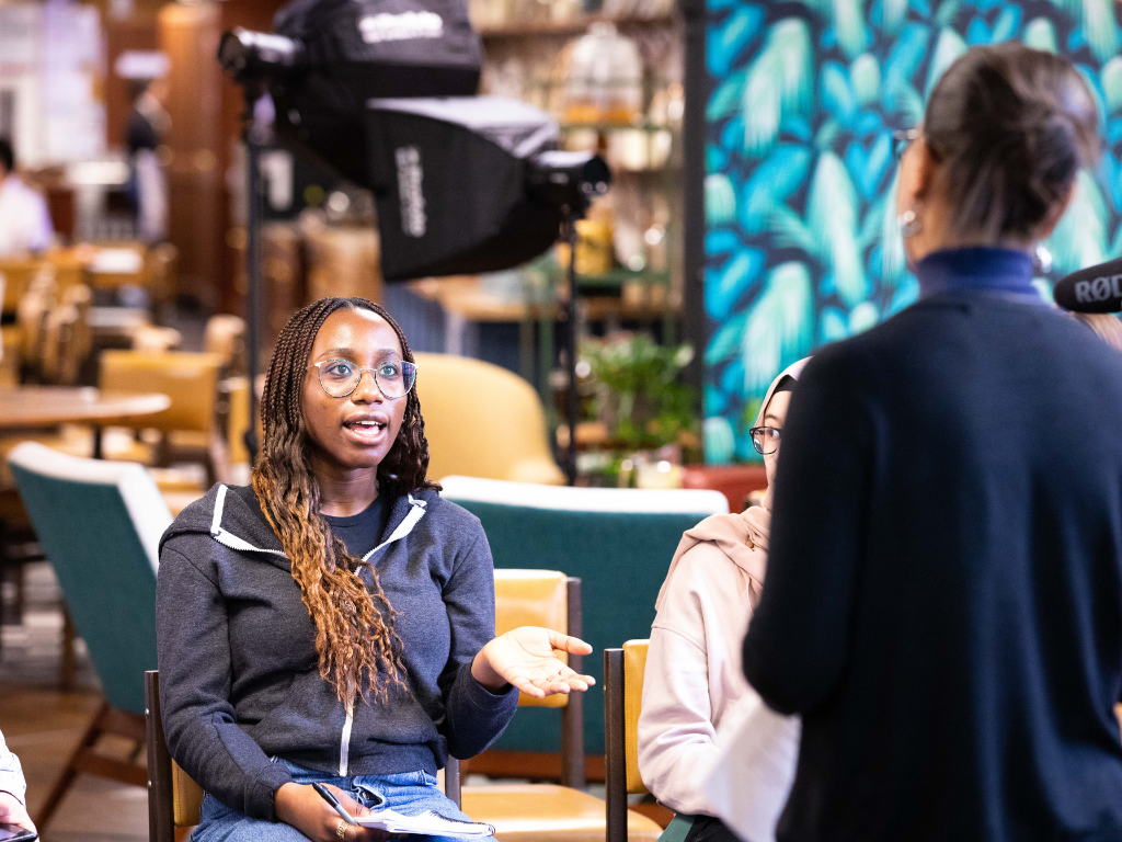 Becky, a young black woman, with long braids that go from dark brown to blonde at the tips, is speaking to a woman who has dark hair in a bun and her back to the camera. Becky is wearing a grey zip hoodie and blue jeans, and is facing the woman while sat down on a chair.