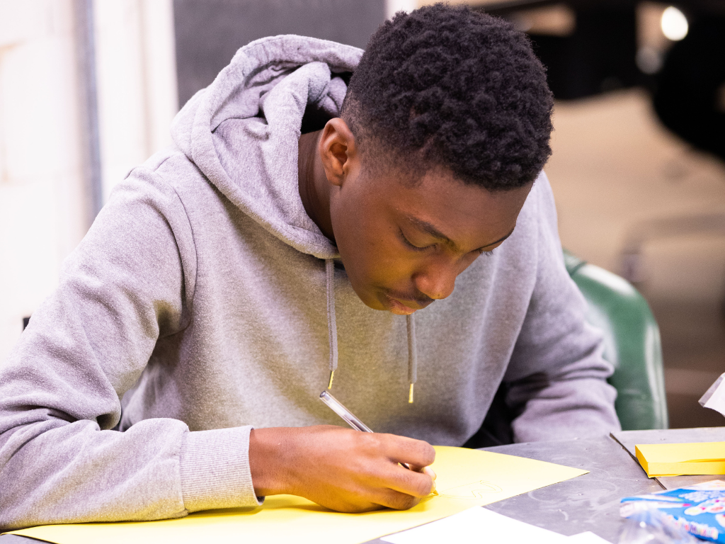 Victor, a young black male in a grey hoodie, is leaning over a table and writing on a yellow sheet of paper.