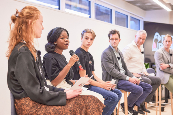 A young black woman is speaking into a mic at a panel event, addressing the room. Next to her is a young white man, both are Bite Back activists wearing black Bite Back shirts. The rest of the panel is watching the speaker.