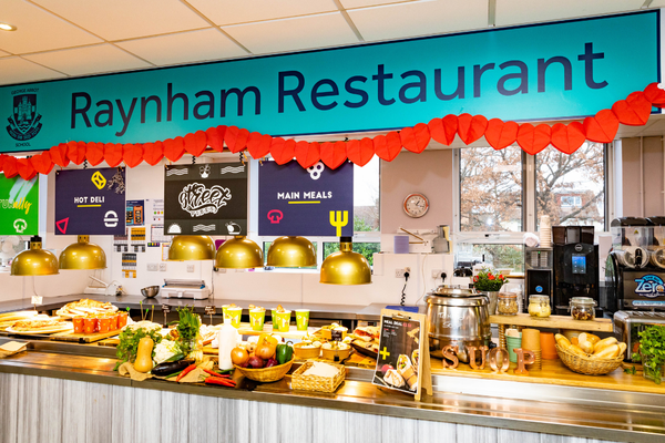 A school dining room, where a counter displays a range of healthy options, such as bread, a large container of soup and fruit. On top there is a banner that states the name as 'Raynham Restaurant' in large navy letters on a turquoise background with red piping at the bottom