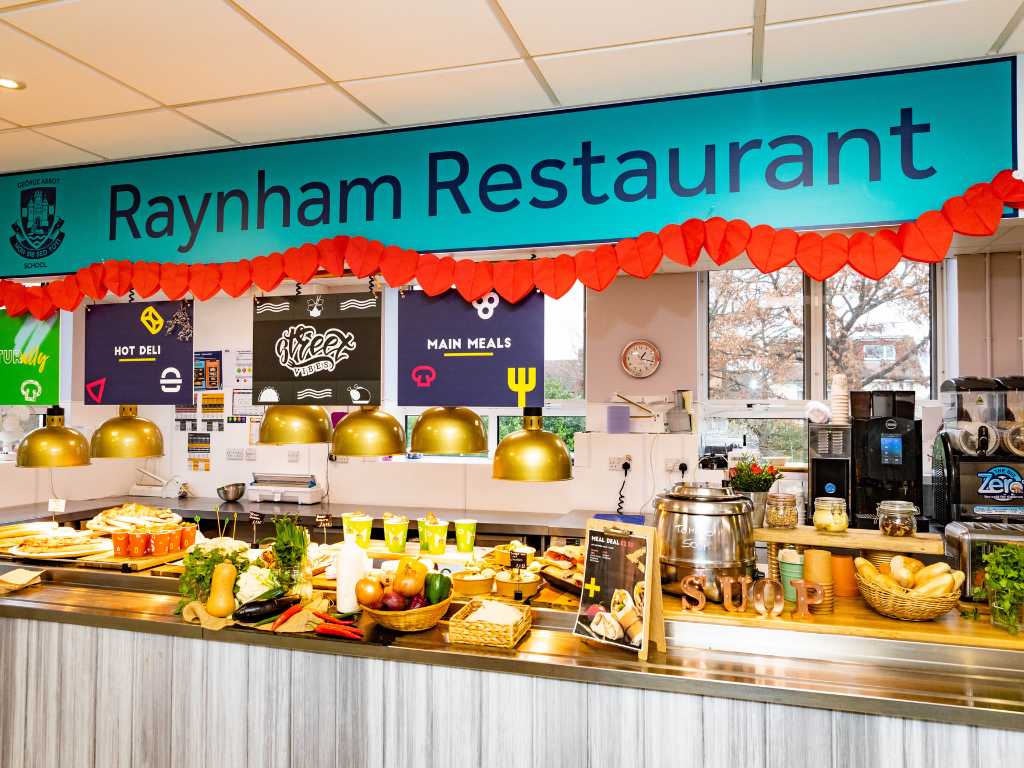A school dining room, where a counter displays a range of healthy options, such as bread, a large container of soup and fruit. On top there is a banner that states the name as 'Raynham Restaurant' in large navy letters on a turquoise background with red piping at the bottom