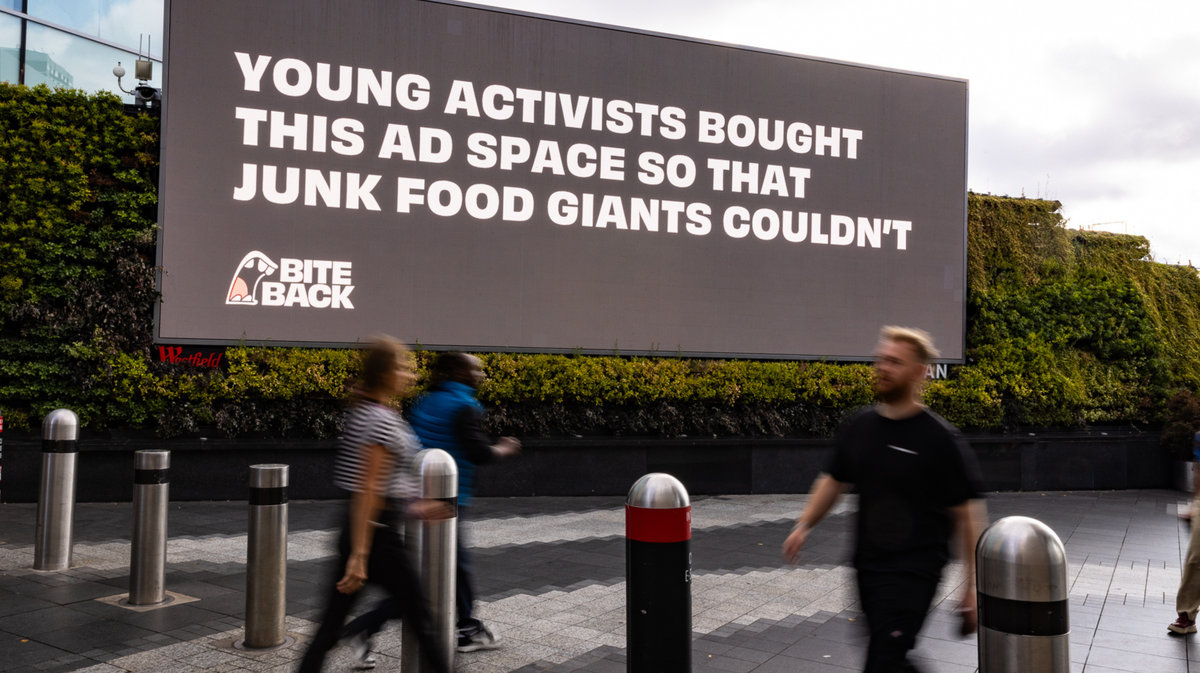 A big black bill board with the writing "Youth activists bought this ad space so junk food giants couldn't" in white with various people passing by in movement in the foreground. Bite Back junk food