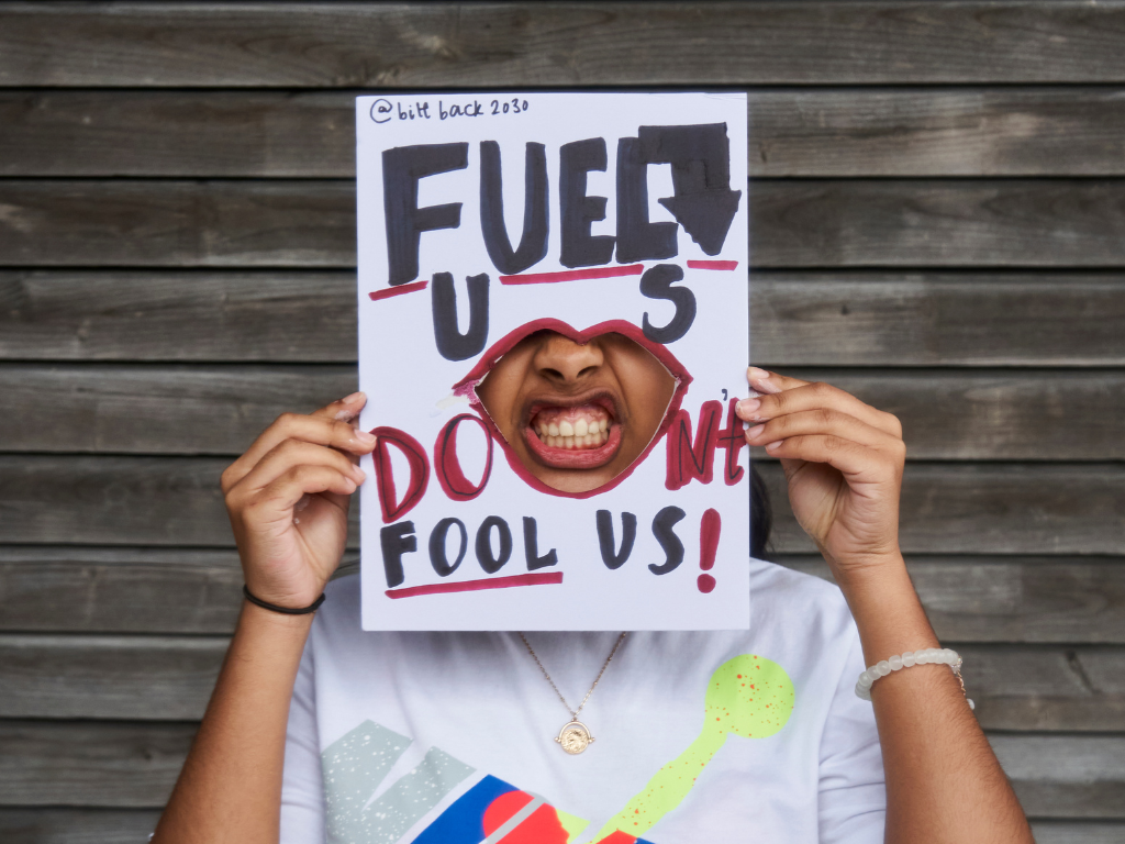 A protest sign saying "Fuel Us Not Fool Us" in red and black lettering with a cut out mouth and a brown skinned activist holding it up and showing their clenched mouth through it. They are standing in front of a dark wooden wall.