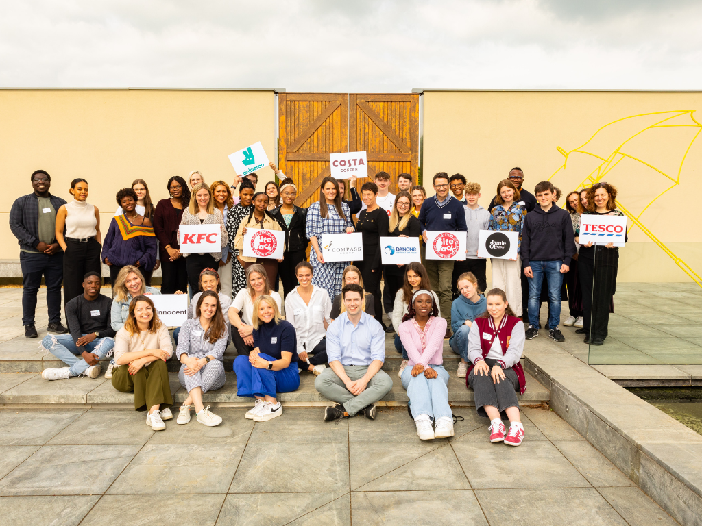 All attendees of the Food System Accelerator launch posing for a photo on the steps outside the venue in front of a yellow wall with a wooden door. They are holding signs signifying their companies (Tesco, Innocent, KFC, Deliveroo, Danone, Compass and Jamie Oliver)