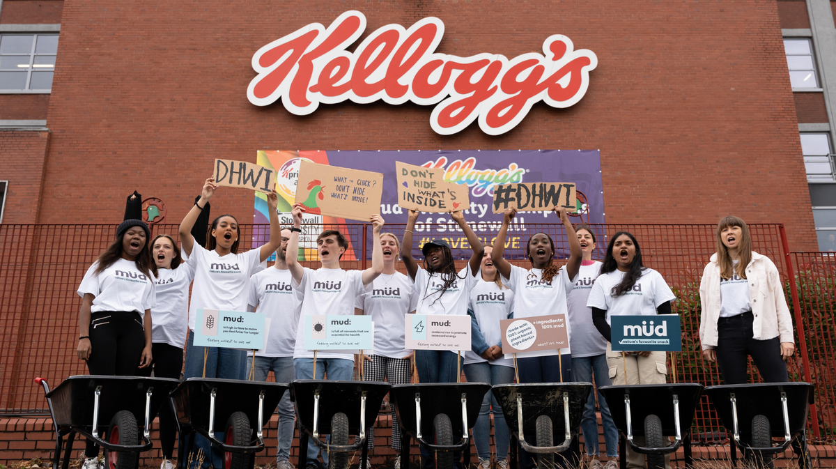 A group of young activists in white t-shirts standing in front of the Kellogg's head office holding signs saying things like #DHWI and Don't Hide What's Inside. The Kellogg's logo with the rooster is framing them from behind.