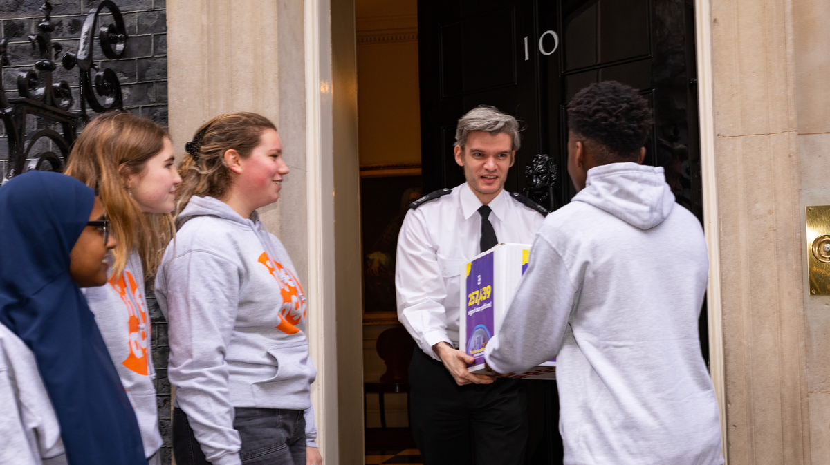 Victor handing over a white box with our FSM petition to a smiling man in uniform with white hair standing in an open doorway with a black door. The door to No.10. Amy a young white girl with red blondish curly hair, Molly a young white girl with straight shoulder length blonde hair and Yumna look o