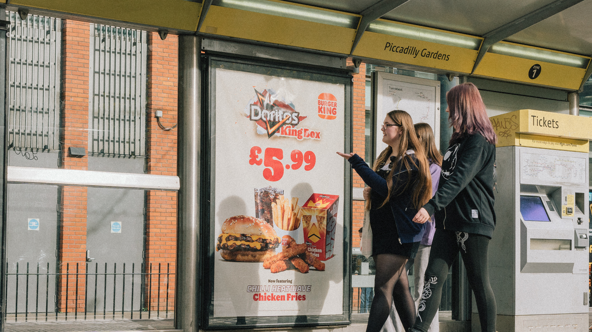 Three activists strolling past a bus stop featuring a large advert for a collaboration between Burger King and Doritos displaying a burger, large soft drinks and chicken fries.