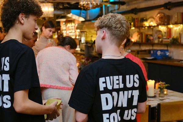 Two young activists Vin on the left and Jacob on the right standing with their back to the camera showing the back of their shirts saying 'Fuel Us Don't Fool Us'. They are standing in a dark, dimly lit event space surrounded by other attendees.