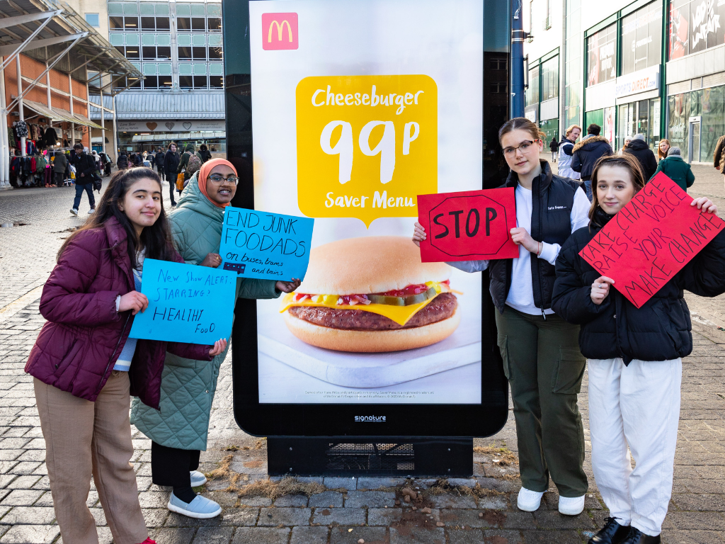 A group of young activists from Birmingham standing next to a McDonald's Saver Menu ad holding signs saying 'Stop & End Junk Food Ads'. The actvists are dressed for the cold in thick coats and long warm trousers.