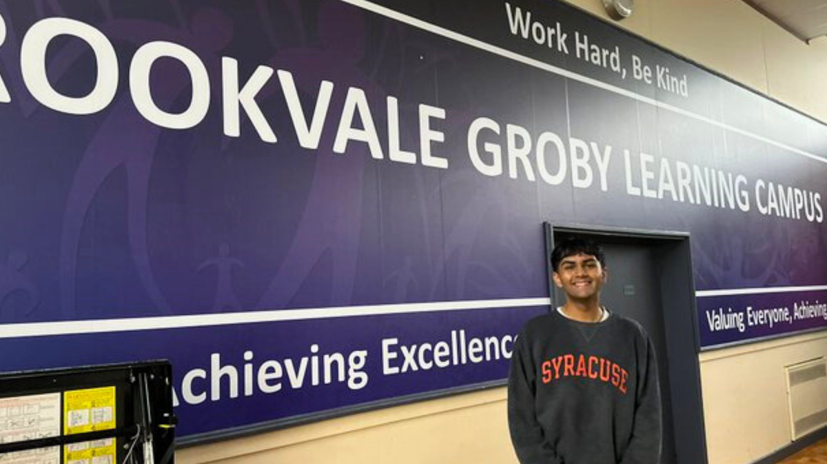 Activist Dev, a young brown man with black hair cut into a bowl cut stands in front of a large blue banner with white writing reading Brookvale Groby Learning Campus. He is wearing a dark grey sweater with the word Syracuse in red writing on it with black jeans and has a big proud smile on his face.