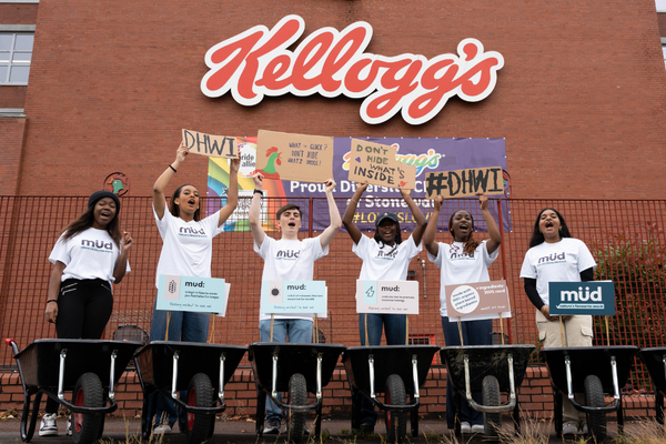 A group of young people standing in front of Kelloggs headquarters with the famous chicken on a red brick building behind them. They are wearing white shorts and have wheelbarrows with mud in front of them.