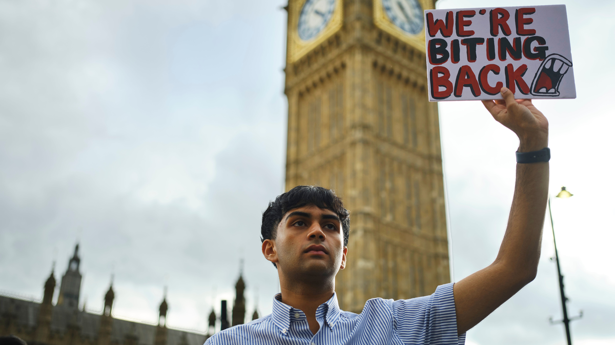 Young activist Dev, a young tanned man with his black hair cut into a bowl cut and wearing a pale blue shirt, is holding up a protest sign saying "We're Biting Back" outside Parliament in September 2023. A tower of the houses of parliament is visible behind him.