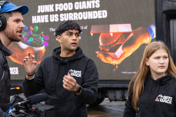 Young activists Dev & Maya are standing on a busy pavement in front of a digivan that displays an image of hands reaching towards a child with burgers and candy and the caption 'Junk Food has us surrounded". They are speaking to a camera man holding a large camera. Dev is a young brown man with his black hair cut into a bowl cut and thick eye brows and Maya is a girl with a very pale complexion and dirty blonde straight hair . Both are wearing dark hoodies that show the iconic Bite Back logo.