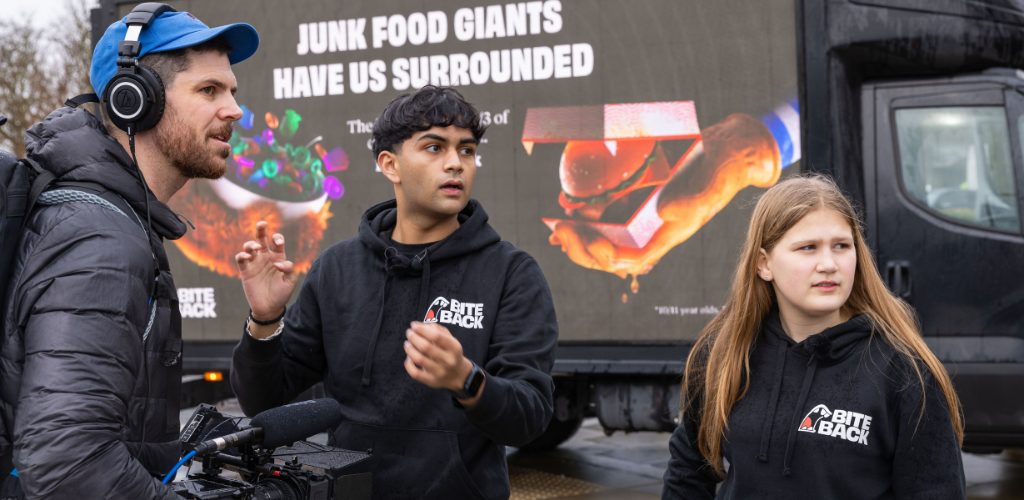 Young activists Dev & Maya are standing on a busy pavement in front of a digivan that displays an image of hands reaching towards a child with burgers and candy and the caption 'Junk Food has us surrounded". They are speaking to a camera man holding a large camera. Dev is a young brown man with his black hair cut into a bowl cut and thick eye brows and Maya is a girl with a very pale complexion and dirty blonde straight hair . Both are wearing dark hoodies that show the iconic Bite Back logo.