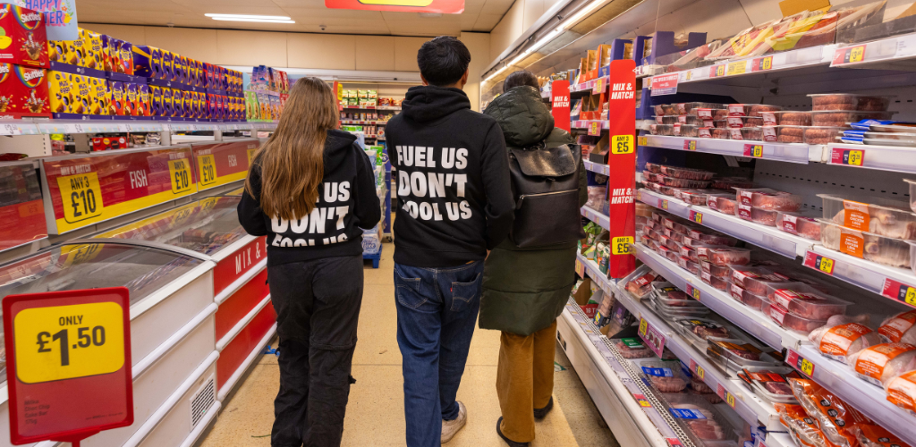 Two young people are walking through a supermarket aisle. On both sides of them is an overwhelming array of brightly coloured products encroaching on them. Both are wearing black hoodies saying "Fuel Us, Don't Fool Us"