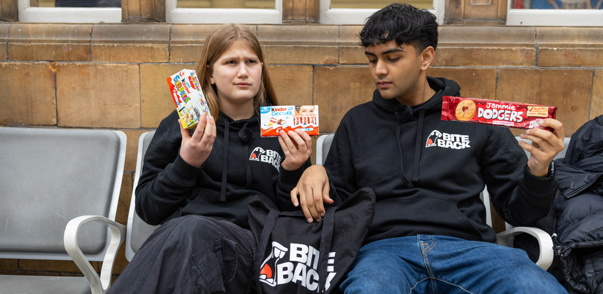 Maya and Dev two Bite Back activists sit on a bench in front of a brick wall. It could be a station platform. Maya and Dev are holding up junk food products such as Kinder chocolate bars and Kinder surprises or Jammie Dodgers. Maya is making a face that suggests she is confused why she is being bombarded by these products and fed up with it.