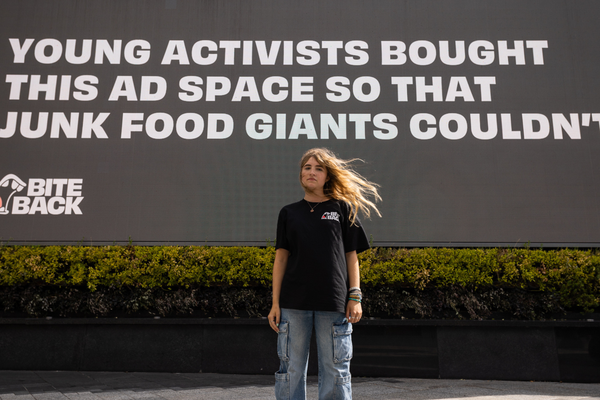 Young activist Molly a young girl with a pale complexion, freckles and long golden hair is standing in front of a large billboard at Westfield White City that reads "Young activists bought this space so that junk food giants couldn't." Molly is wearing a serious determined expression showing she is ready to fight back against a food system set up to fail us all.