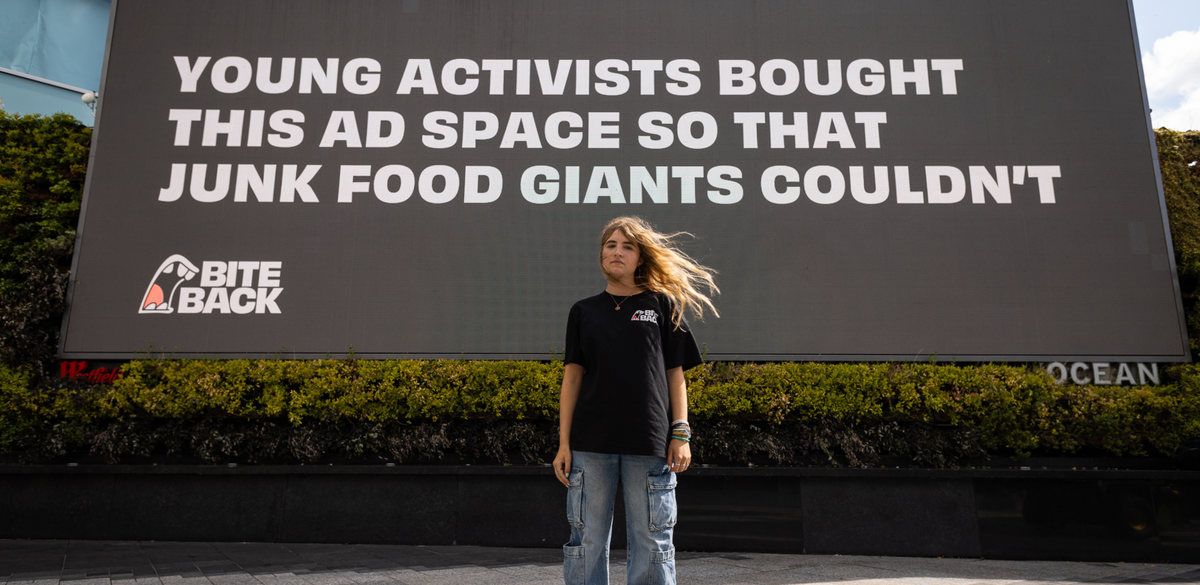 Young activist Molly a young girl with a pale complexion, freckles and long golden hair is standing in front of a large billboard at Westfield White City that reads "Young activists bought this space so that junk food giants couldn't." Molly is wearing a serious determined expression showing she is ready to fight back against a food system set up to fail us all.