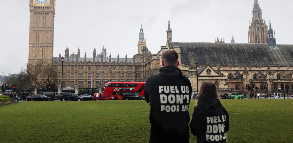 2 young people with their backs to the camera are facing Westminster building. They are wearing black Bite Back hoodies which have white text on the back that says, 'Fuel us, Don't fool us'