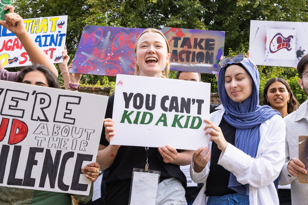 A group of Bite Back activists stand outside holding their activist art, including protest style signs and a paper megaphone. In the centre of the image activist Clemmie is smiling while holding a sign that reads You Can't Kid A Kid