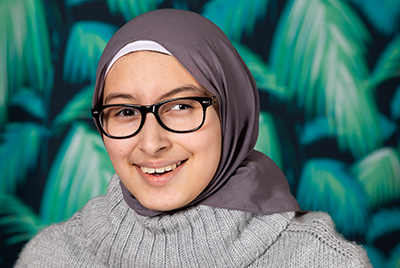 Zainab is a young girl wearing a grey headscarf tucked into a grey turtleneck jumper. She wears round black glasses on her face and is smiling at the camera, while stood in front of green, leafy wallpaper.